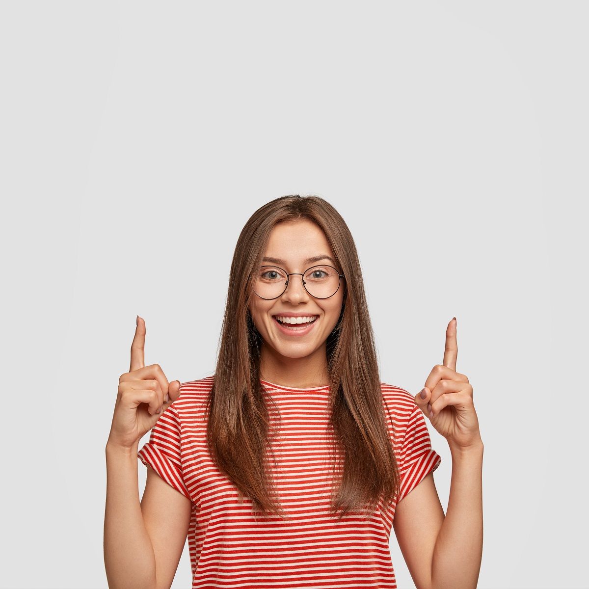 Dreamy positive woman with satisfied expression, points with both index fingers upwards, thinks about new possibilities, shows blank space for your advertisement, stands indoor. Look, big discounts