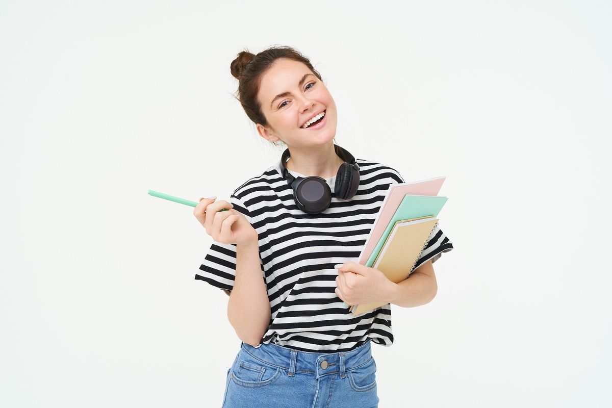 Portrait of young woman, student with notebooks and earphones on her neck, posing for college advertisement, white background